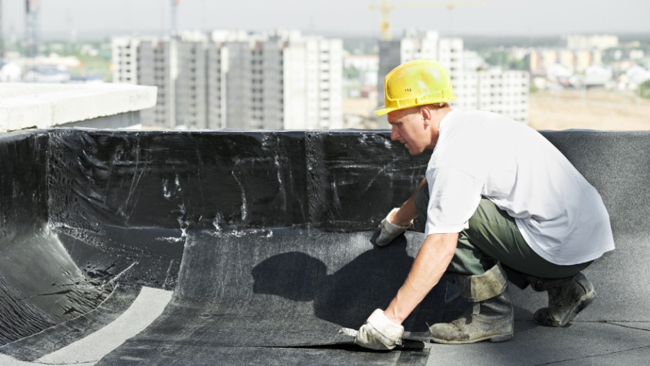 A construction worker in a yellow hard hat installs waterproofing membrane on a rooftop, with tall buildings and cranes visible in the blurred background—ideal work for an emergency roofing contractor in Fullerton skilled in roof tarping.