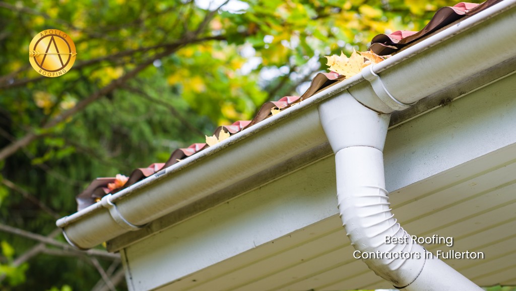 Close-up of a house’s white gutter filled with yellow leaves, highlighting the need for gutter control. Green trees form the backdrop. Text reads Best Roofing Contractors in Fullerton with the company logo in the top left corner.
