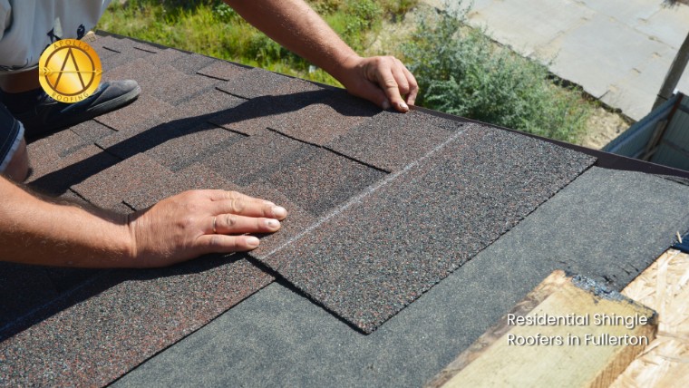 A person installs asphalt shingles on a rooftop, carefully aligning them to help prevent shingle roof granule loss. Text reads "Residential Shingle Roofers in Fullerton." A round logo appears in the upper left corner.